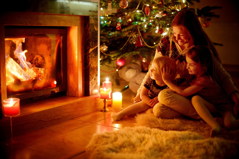 Young mother and her two little daughters sitting by a fireplace in a cozy dark living room on Christmas eve