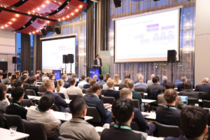 A large group of people in a conference hall, watching a presentation held by a person on a stage.