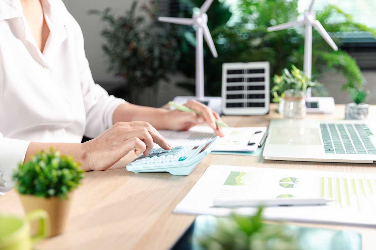 A person at a calculator, with miniatures of wind turbines and solar panels in view, as well as a laptop.